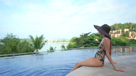 asian woman in swimsuit sitting on pool edge and contemplating panorama