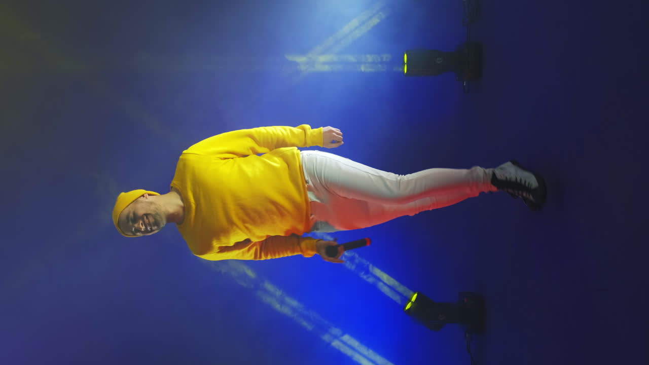 video vertical. un cantante con un traje amarillo baila y canta con un micrófono de color neón. salta y mueve vigorosamente.