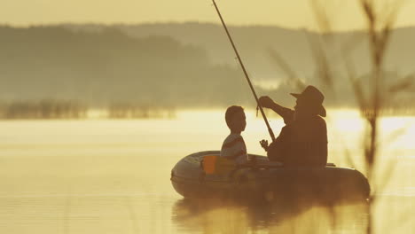 vista trasera de un anciano con sombrero y su nieto en un barco pescando con una caña en el lago en una mañana nublada