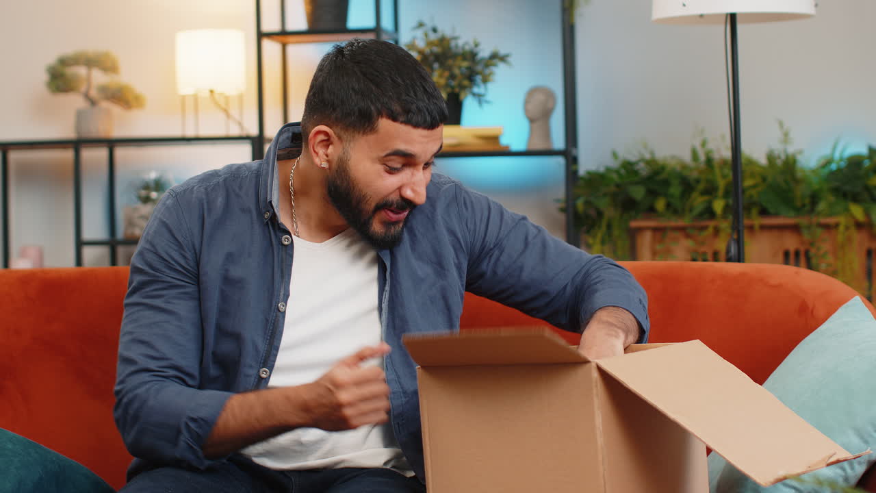 hombre indio desempaquetando paquete de entrega recibiendo compra por correo postal mensajero sentado en el sofá