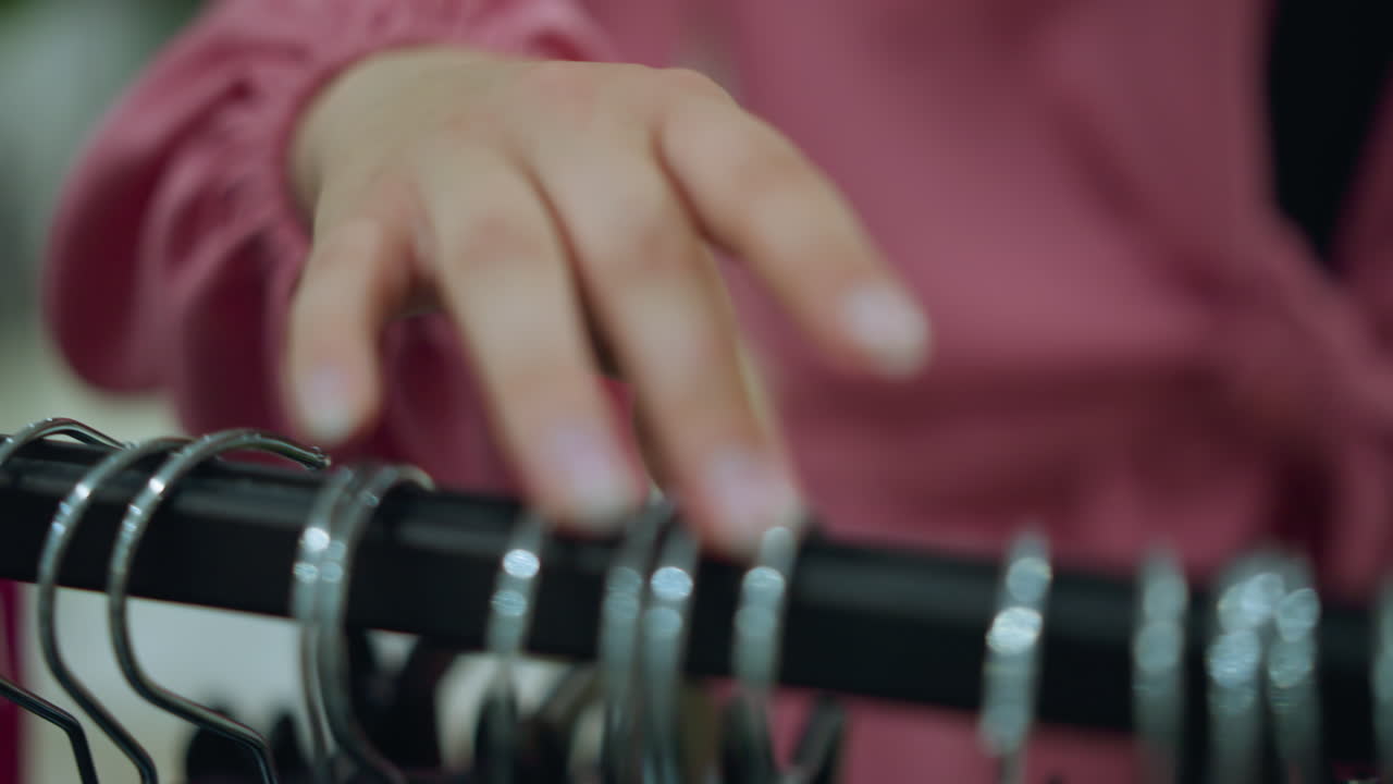 Hand in a pink shirt browsing through a clothes rack filled with metal hangers in a brightly lit mall as the person stops to pick from the rack, with blur view of other items in the background