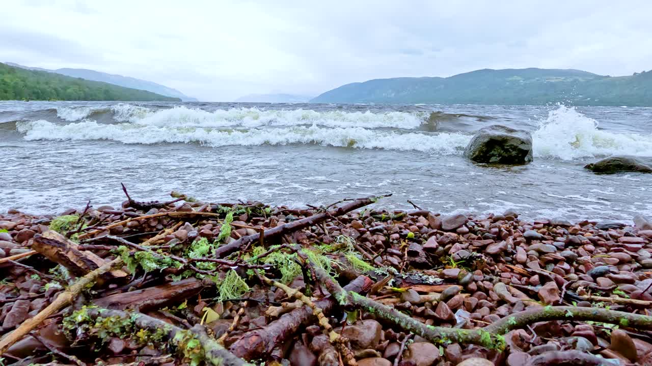 Low-angle view of waves rolling onto a pebble and driftwood-strewn shore at Loch Ness, under overcast daylight, with gentle camera movement