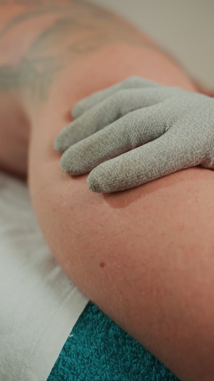 Primo piano di un braccio caucasico che riceve un'esfoliazione con guanto testurizzato, estetista che esegue un massaggio a microcorrente sul braccio superiore tatuato su un asciugamano color ottanio in un ambiente spa calmo, granuli di sale e dettaglio della pelle.