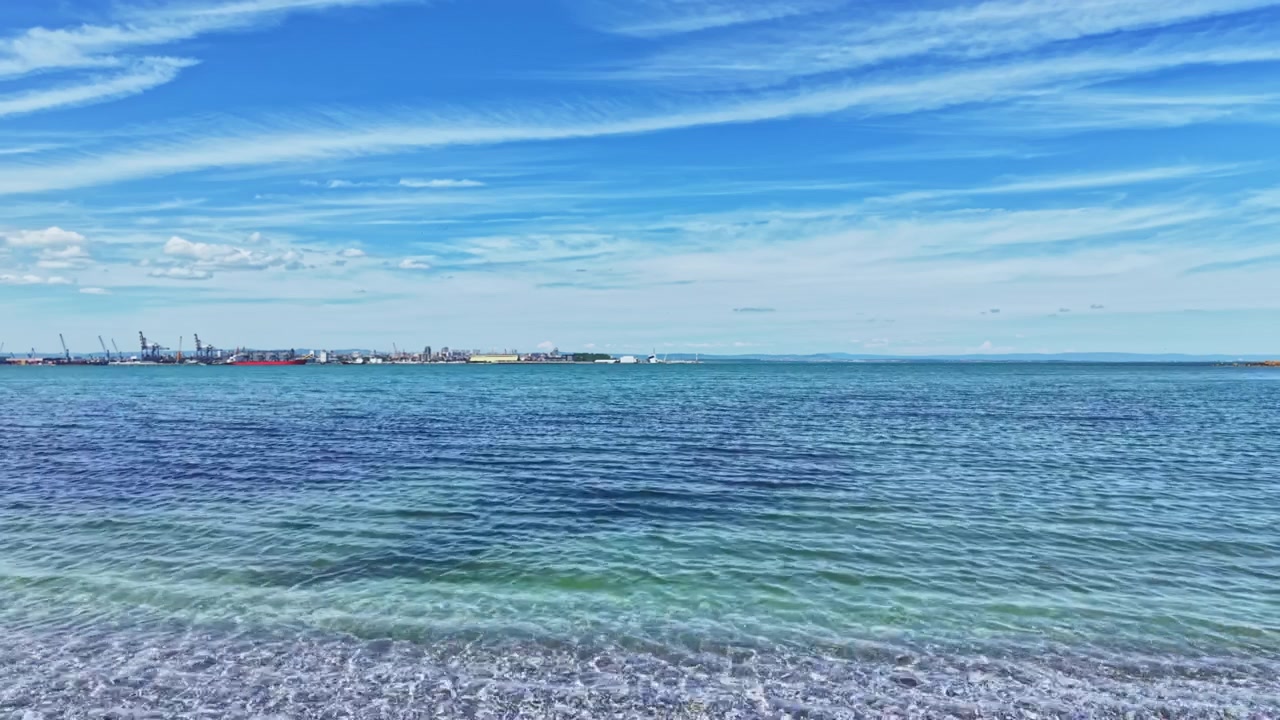 Ruhiger azurblauer Ozean, der sich bis zum Horizont erstreckt, mit sanften Sonnenlichtreflexionen. Eine stille und friedliche Ozeanszene mit sanfter Brandung, die Kieselsteine unter einem leicht bewölkten blauen Himmel berührt.