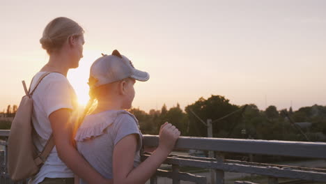 a woman with her daughter are standing at the railing of the bridge at sunset evening walk with mom