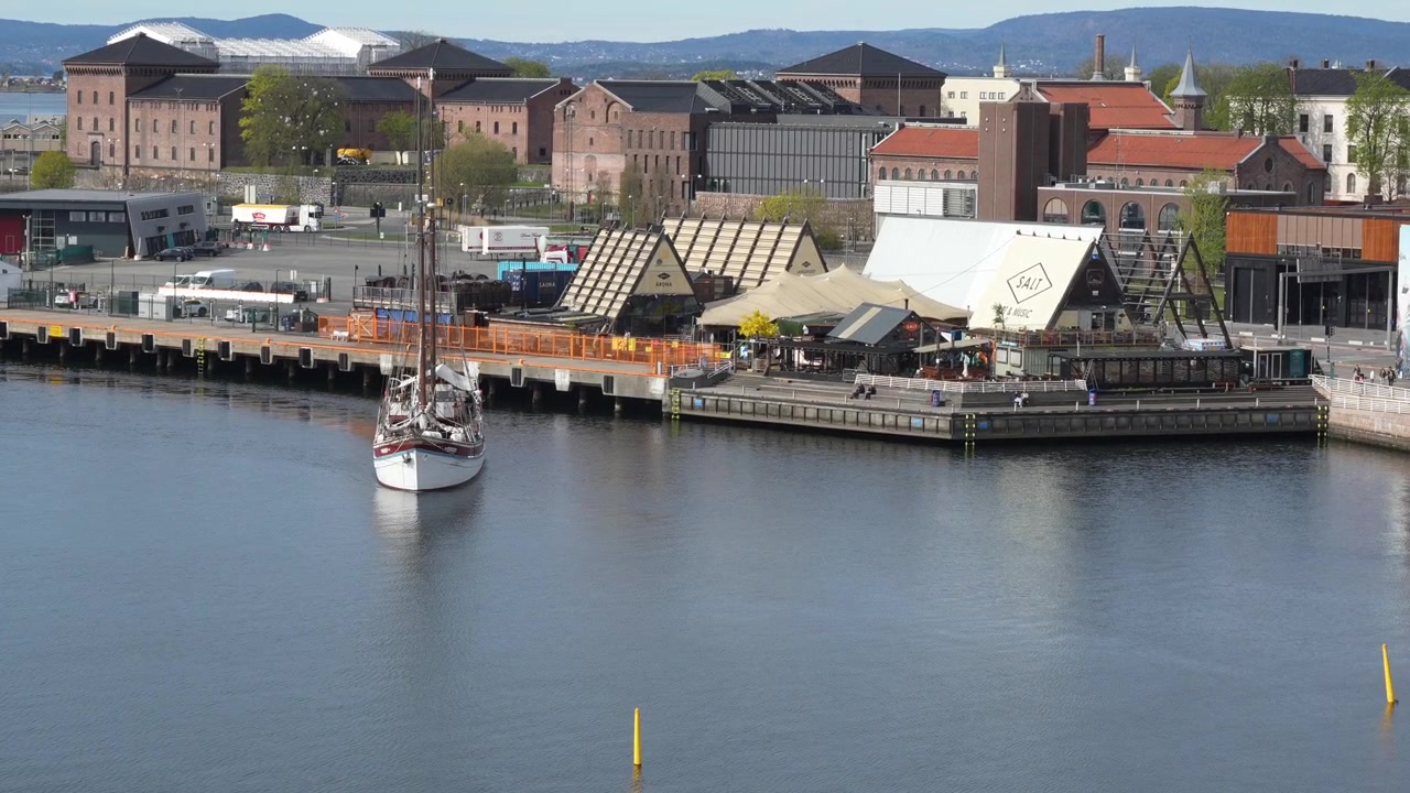 Plano general a mano alzada con vista a un barco turístico de madera deslizándose por el fiordo de Oslo junto a vibrantes edificios ribereños, capturado a plena luz del día desde la azotea de la Ópera de Noruega