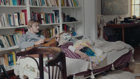 couple resting on bed at home, boy playing the guitar to girlfriend
