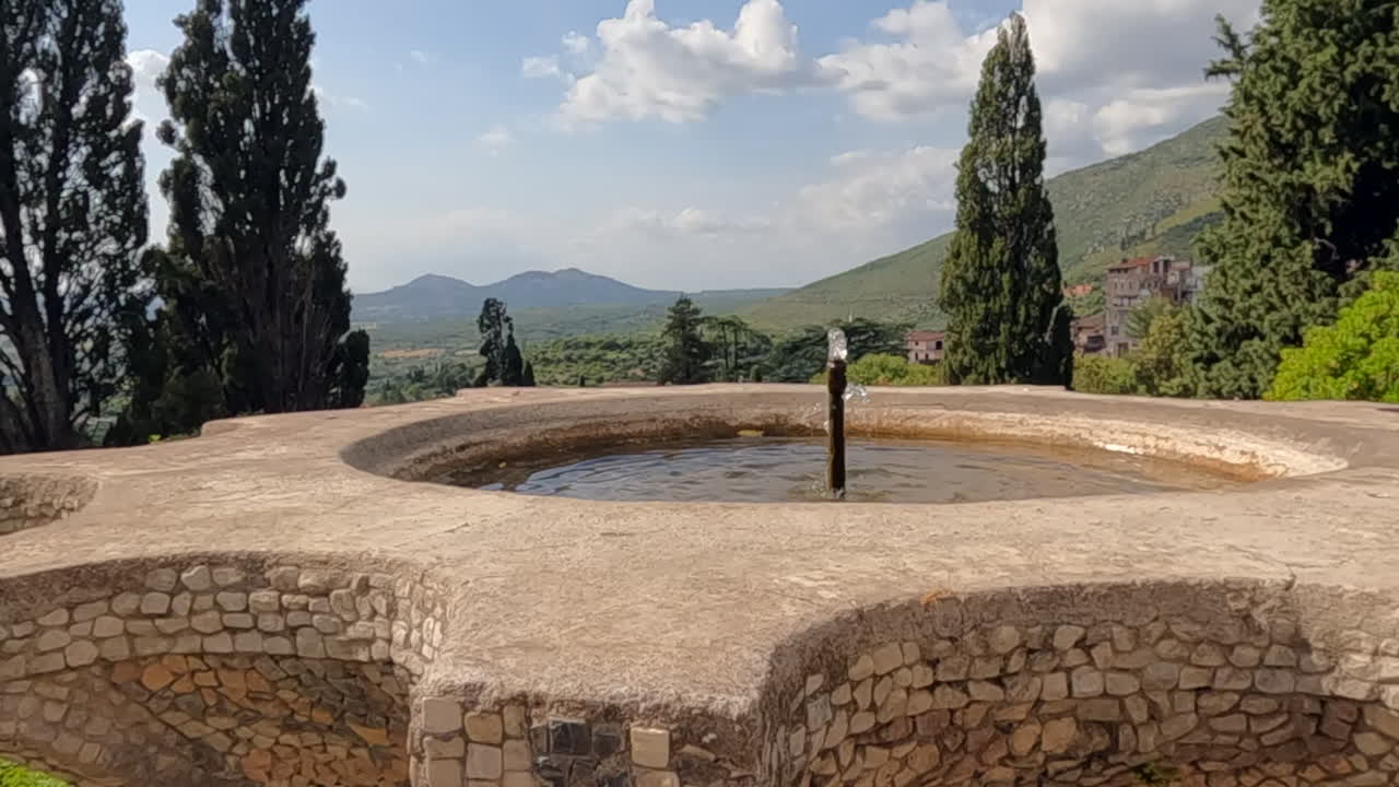 Storica fontana in pietra a Tivoli, Italia, con viste panoramiche sulle montagne, cipressi e cielo azzurro. Classico paesaggio della campagna italiana perfetto per contenuti di viaggio e cultura.
