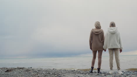 Mother-and-daughter-look-at-the-ocean,-where-storm-clouds-are-in-the-distance.-Standing-on-the-shore,-holding-hands