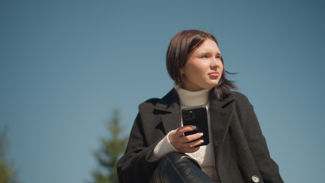 흑색 코트와 색 스웨터를 입은 젊은 여성이 밖에서 벤치에 앉아 스마트폰을 손에 들고 멀리 바라보고 있으며, 밝은 빛이 그녀의 얼굴과 녹색 배경을 비추고 있습니다.