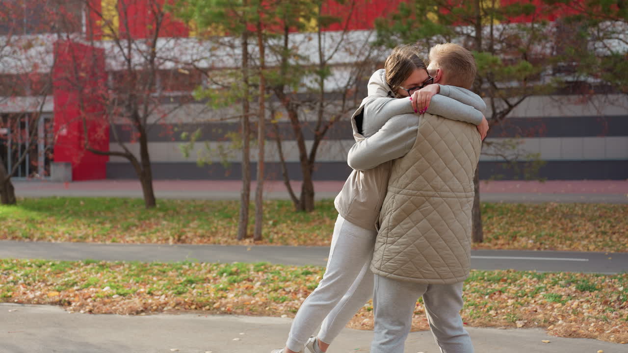 Lady rushes joyfully to hug brother who lifts her and spins her around with love as boy runs in background under autumn trees swaying gently in wind and fallen dry leaves cover ground