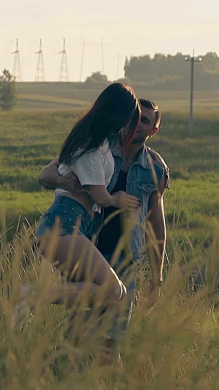 handsome man lit by sun spins young woman with arm against green meadow and forest on horizon slow motion