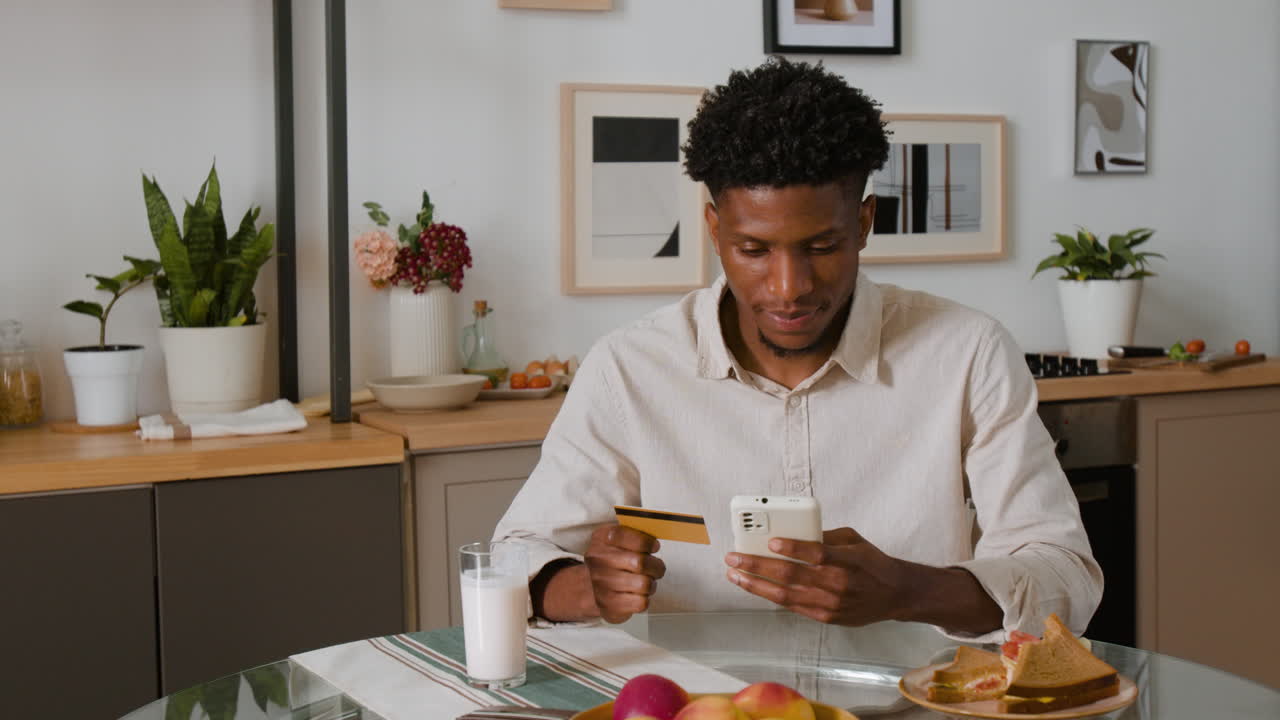Man using smartphone with credit card at kitchen table