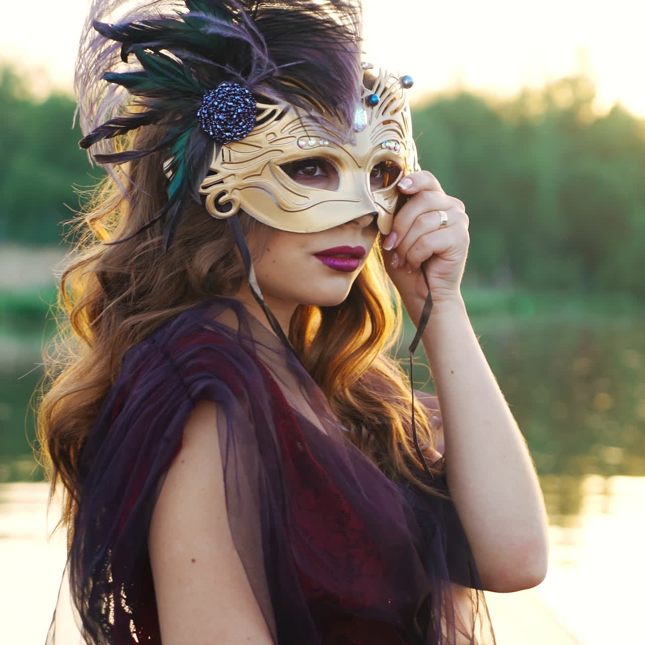 Luxury woman removing carnival mask from her face and looking at powerboat floating nearby at sunset. Beautiful lady in dress holding mask on face standing over the water. Close-up