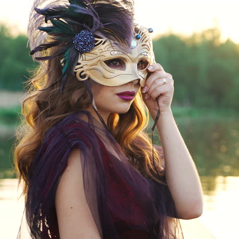 luxury woman removing carnival mask from her face and looking at powerboat floating nearby at sunset. beautiful lady in dress holding mask on face standing over the water. close-up