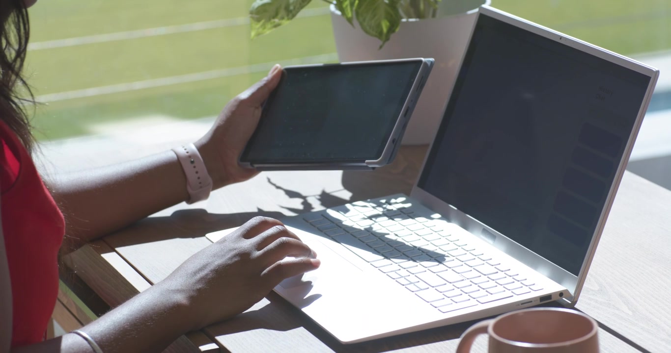 Donna africana adulta che solleva il tablet, tocca e digita sul laptop, lavora vicino alla finestra, con una maglia rossa.
