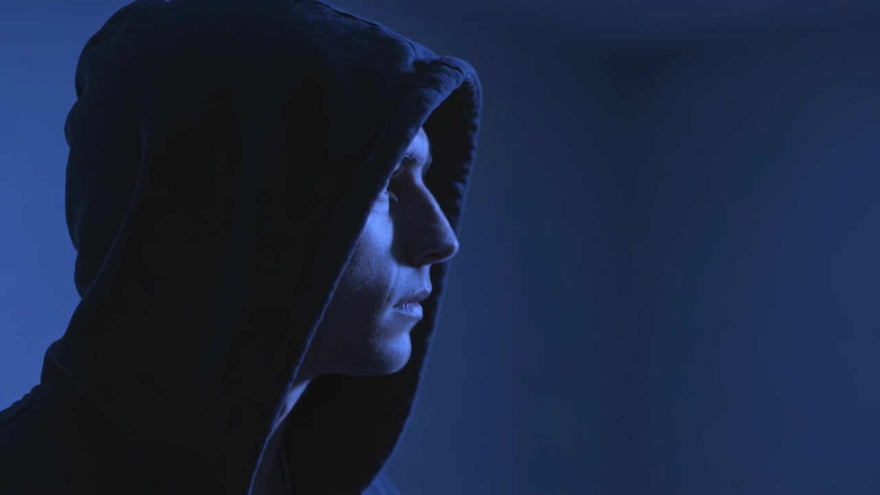 Turning hooded man after lowered gaze revealing left profile at dim blue-lit interior, copy space