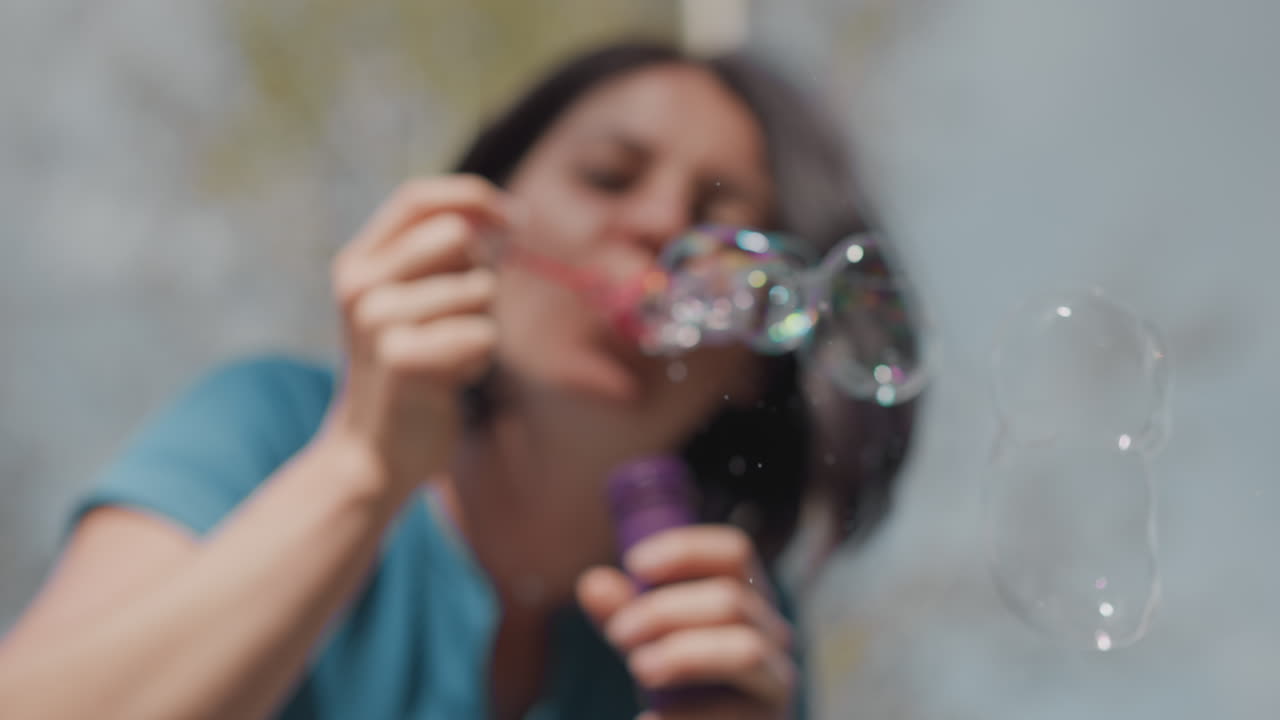 Donna che soffia bolle di sapone alla luce del sole, spensierato momento al parco con sfere iridescenti su sfondo di cielo blu, espressione sorridente e capelli fluenti, flacone di sapone e bacchetta in mano, brezza e movimento energico