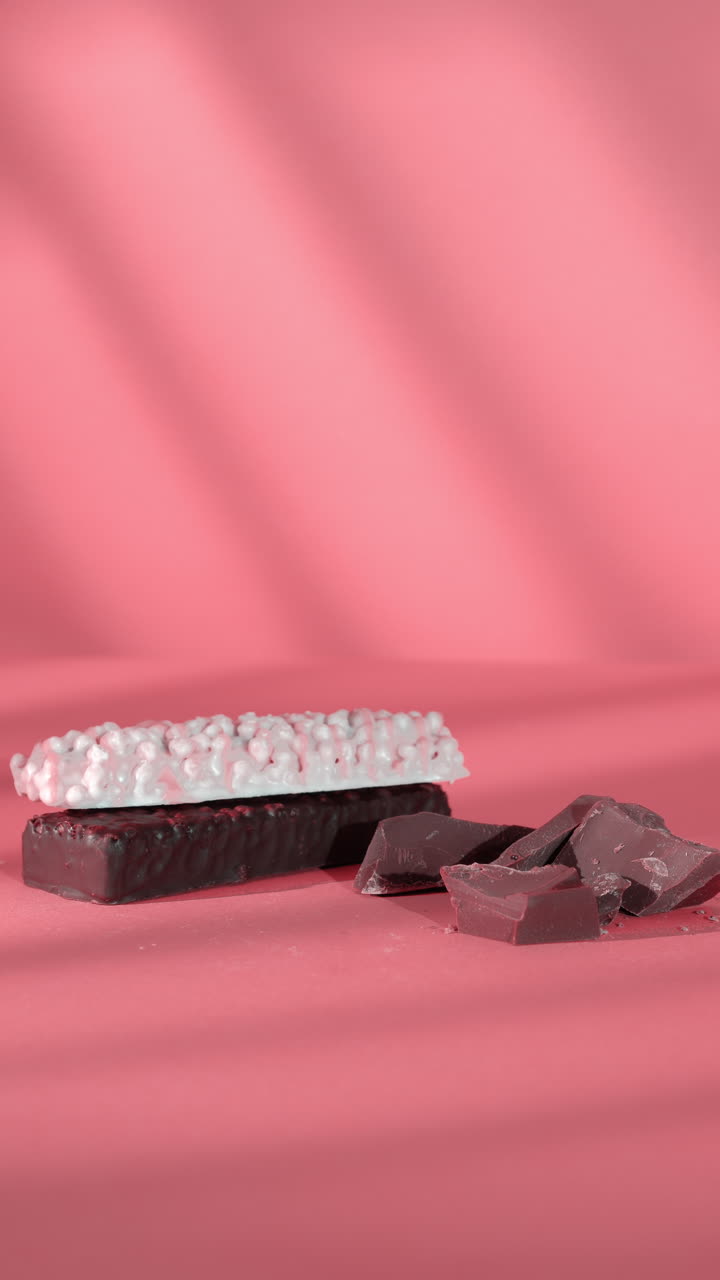 A close-up of a woman's hand delicately placing a puffed white-coated protein bar on top of a dark chocolate bar beside broken chunks of milk chocolate, set against coral pink background soft shadows