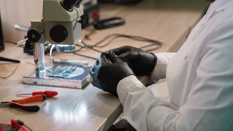 vista de cerca de alguien con una bata de laboratorio ajustando un tornillo en su mano con un componente electrónico bajo un microscopio y varias herramientas en la mesa