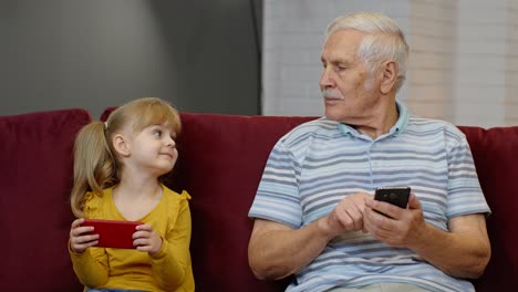 Abuelo-Mayor-Con-Nieta-Niña-Usando-Teléfono-Móvil-Digital,-Jugando-Juegos-En-Casa