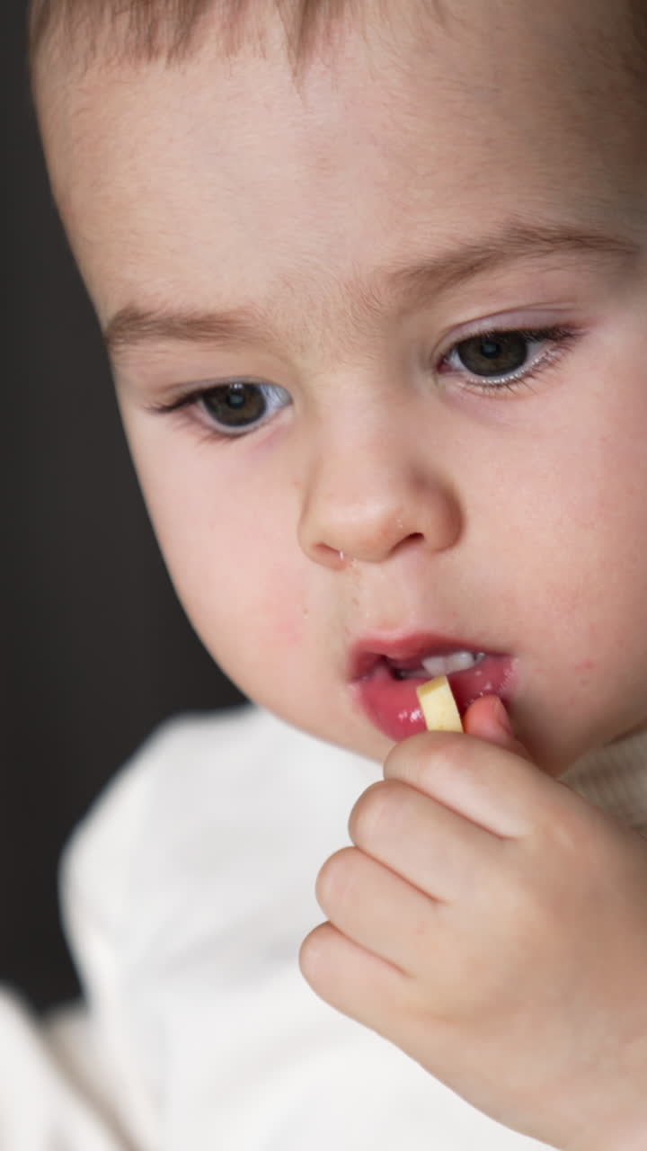 Lovely baby in white sweatshirt takes a piece of cheese from the table. Kid taking food to his mouth thoughtfully. Close up. Vertical video