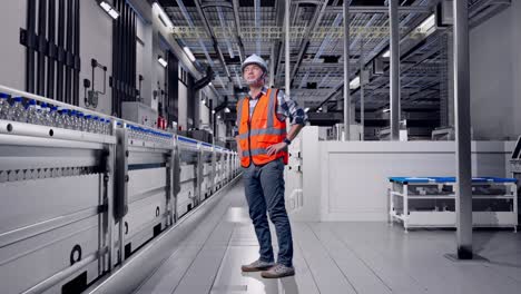 full body side view of asian male engineer wearing safety helmet looking around while standing with arms akimbo with water production in bottling factory