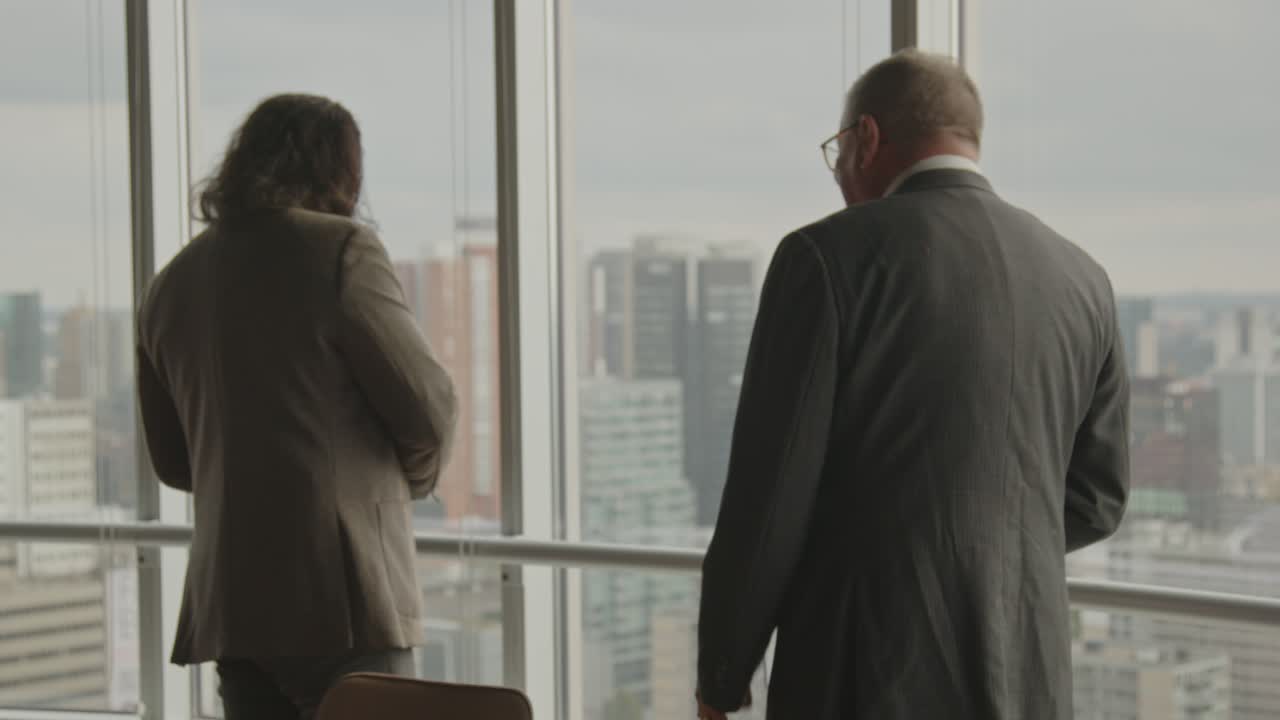 Un CEO anziano parla con rabbia a uno dei suoi dipendenti distratti mentre si trovano davanti a una finestra che si affaccia su uno skyline cittadino.