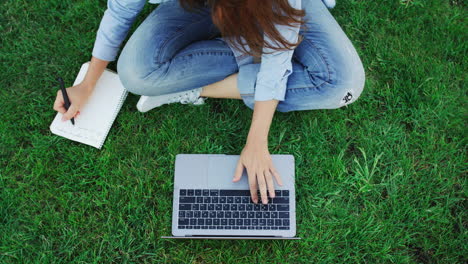 Woman-student-working-on-laptop-computer-and-writing-notes-in-notebook-in-park