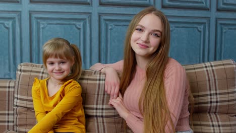 child kid daughter with her young mother hugging and looking at camera while sitting on sofa at home