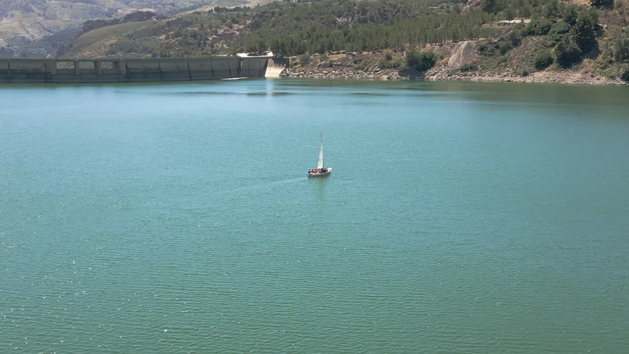 Sailboat sailing next to a reservoir dam on a sunny day. Aerial view in orbit. Water sports. Andalusia. Spain.