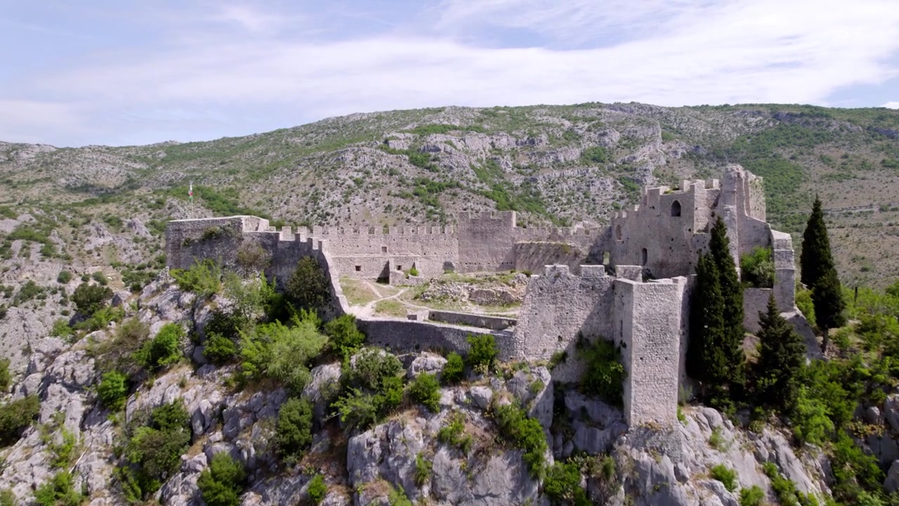 La Fortezza di Blagaj si erge tra rocce calcaree e alberi sopra il paesaggio dell'Erzegovina, sorvolo aereo sopra rovine e mura verso le montagne, offrendo una panoramica