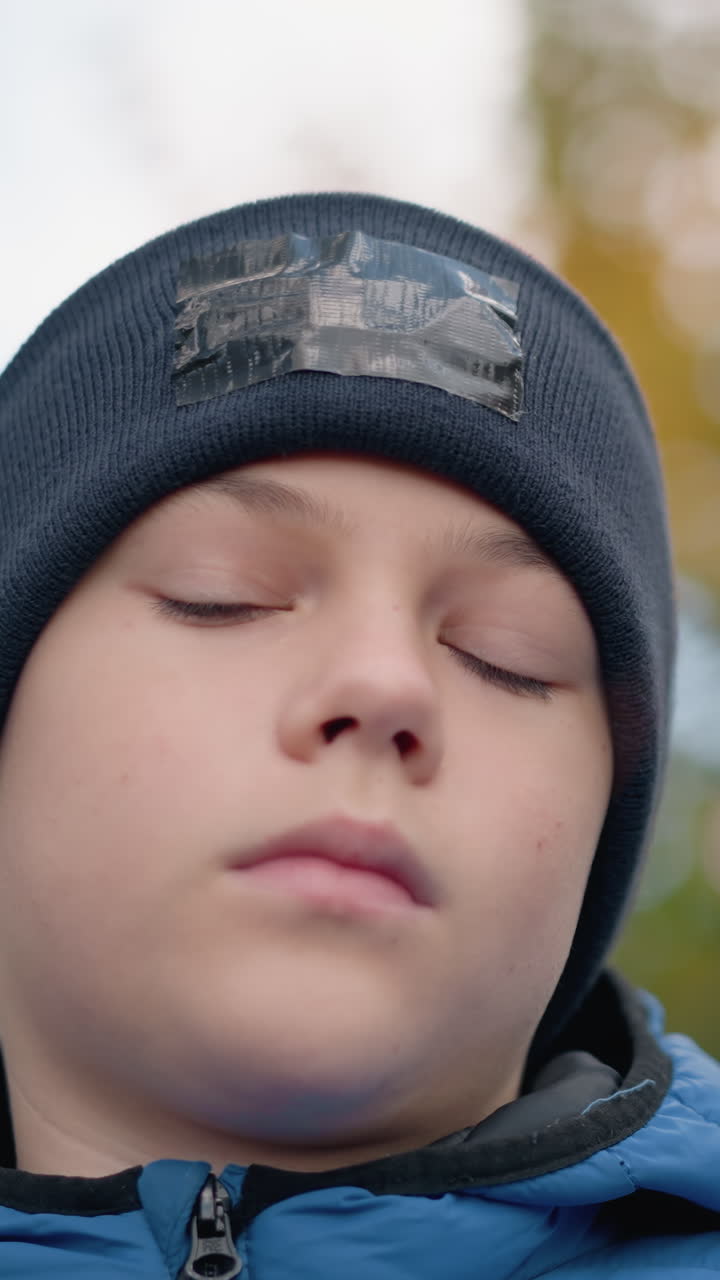 primer plano de un niño con gorra negra y chaqueta azul observando pensativamente una hoja seca antes de dejarla caer, de pie en medio de una vegetación vibrante, fondo borroso con colorido follaje de otoño