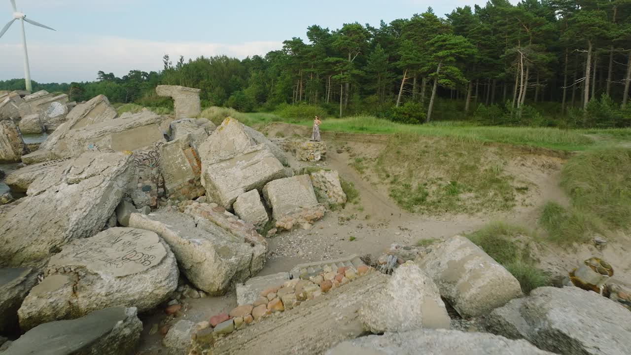 воздушная установка вид красивой молодой романтической кавказской девушки в длинном платье на белом песчаном пляже со старыми руинами, солнечный летний вечер, закат, золотой час, широкий дрон выстрел движется назад