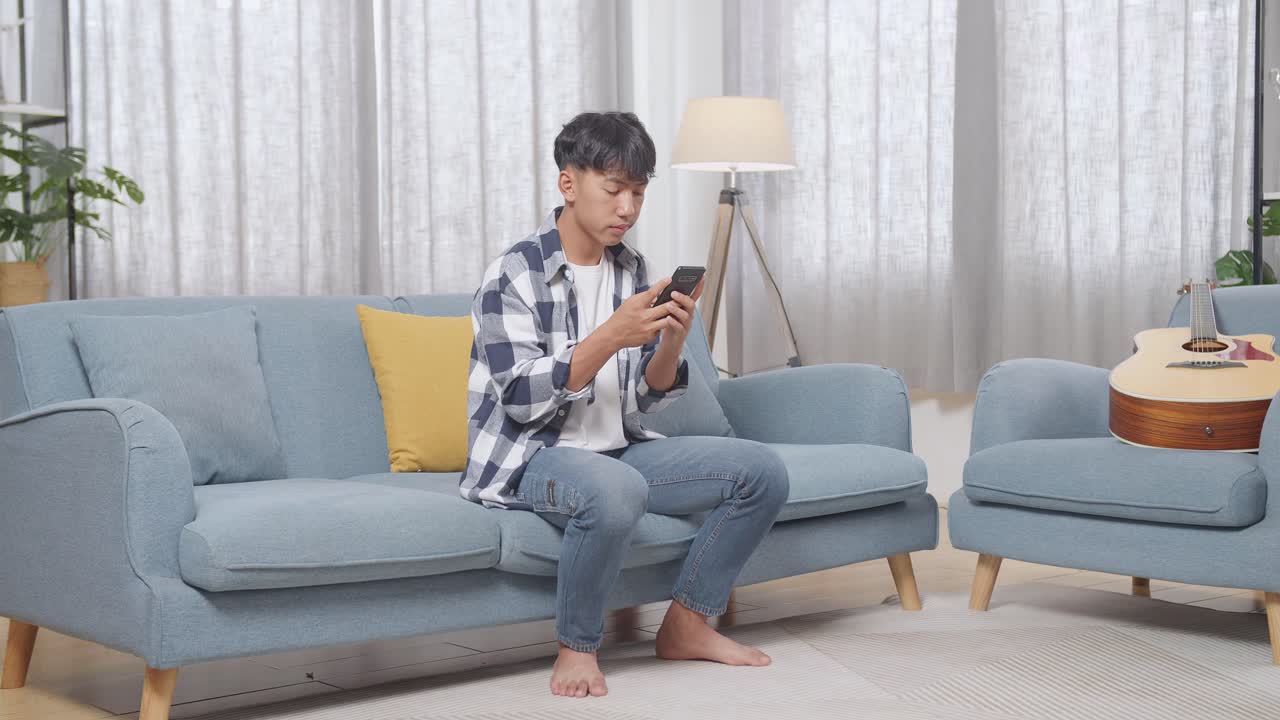 집에서 거실에서 소파에 앉아 스마트폰을 사용하는 지루한 아시아 십대 소년의 몸이 가득 차 있습니다.