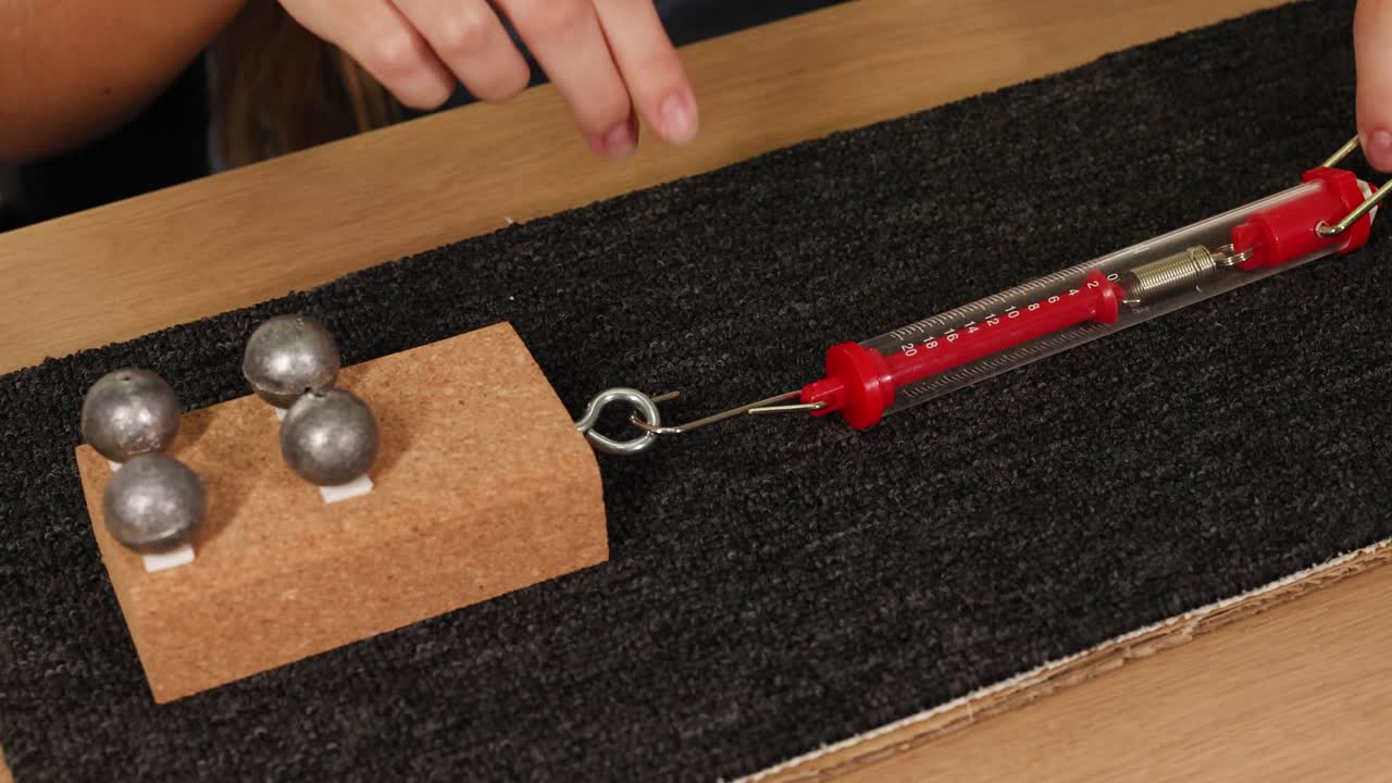 Hands use a spring scale to measure friction on a carpeted surface with weights on a cardboard block