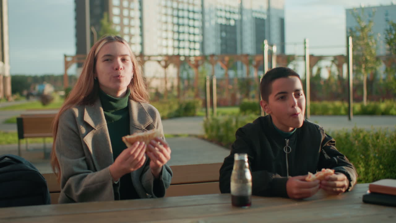 Mum and son sitting outdoors at wooden table in urban park making toast with bread in hand while enjoying snack together, smiling and sharing relaxed family bonding