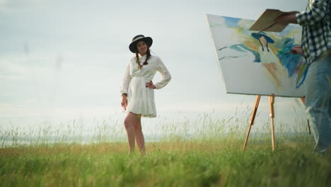 a woman in a white dress and hat poses gracefully in a serene grassy field as an unseen artist in a checkered shirt and jeans paints her portrait on a large canvas