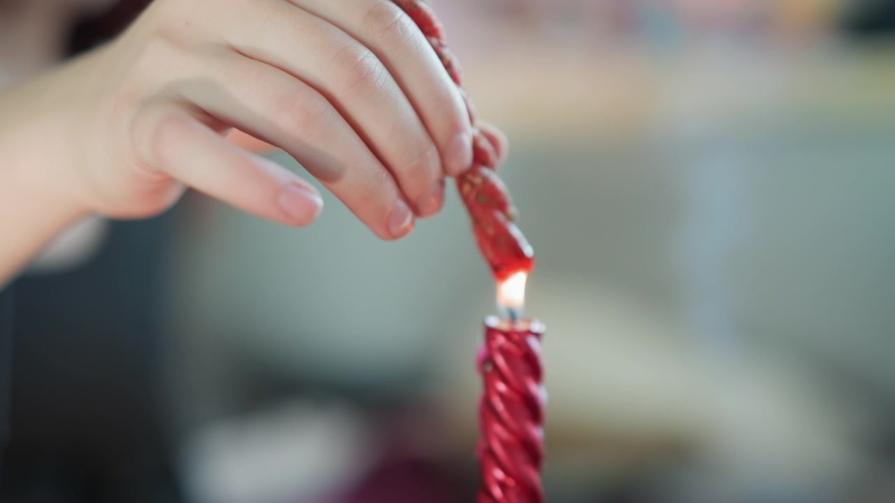 Primo piano delle mani che accendono una candela rossa mentre una donna bianca carica l'intenzione durante un intimo rituale domestico, goccia di cera attorcigliata, fiamma calda, sfondo bokeh soffuso, dettaglio al rallentatore e atmosfera accogliente.