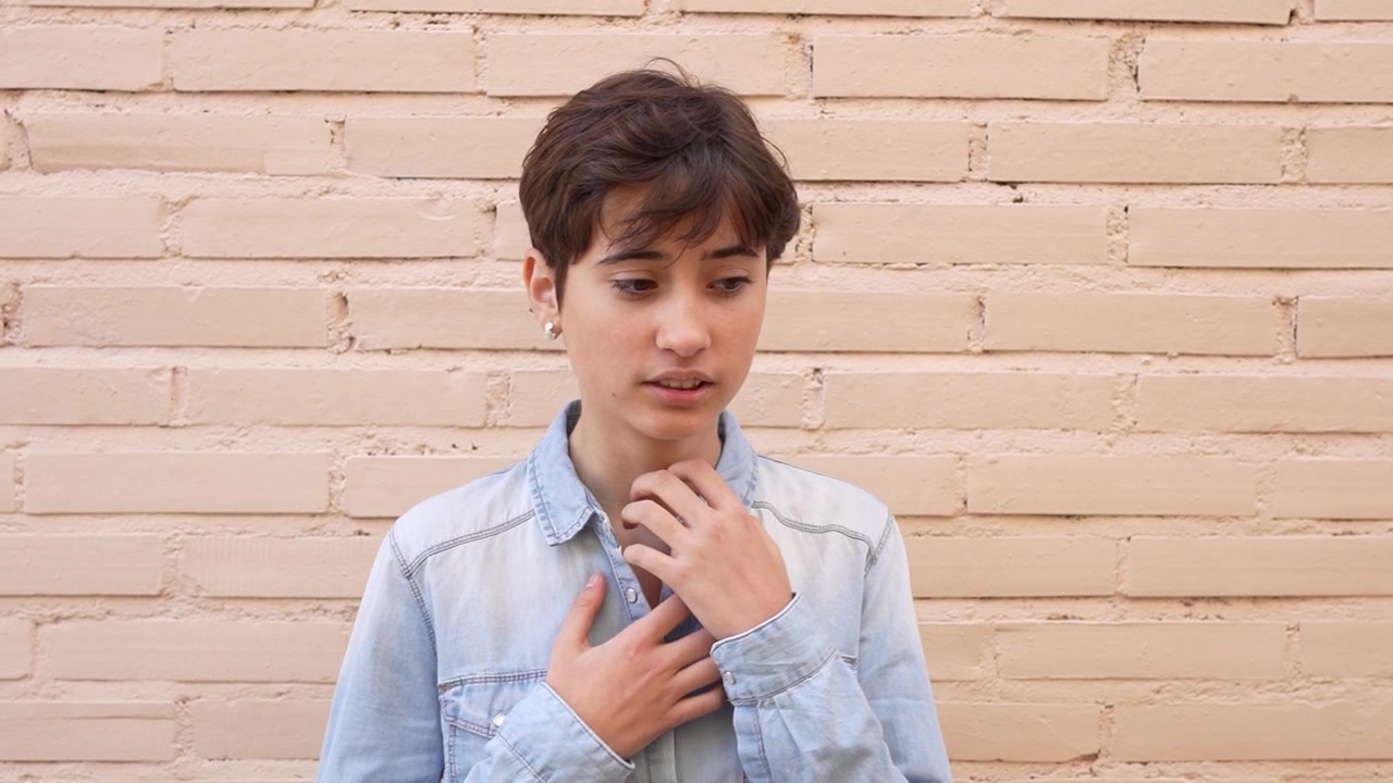 Una giovane persona pensierosa che indossa una camicia di jeans casual in posa contro uno sfondo di muro di mattoni