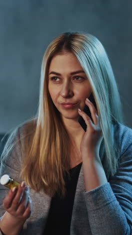 doubting blonde woman calls on smartphone holding small pill bottle against workplace with glowing lamp in semi-dark room close view slow motion