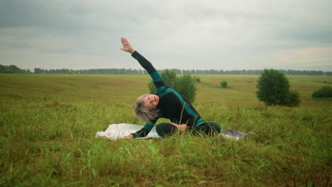 mujer avanzada acostada en un lado de la alfombra de yoga, practicando la postura de flexión lateral con el brazo extendido, sentada en un vasto campo de hierba bajo cielos nublados, rodeada de naturaleza y árboles en la distancia
