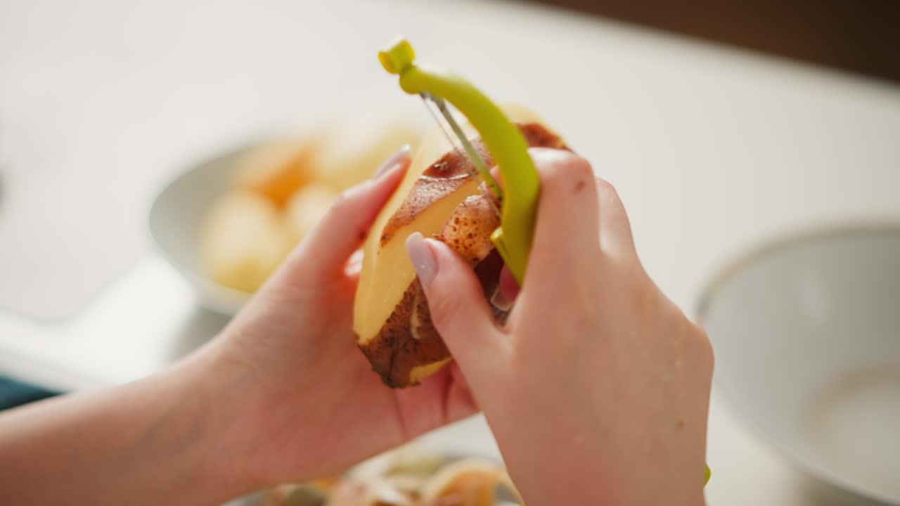 Close up hand view of person peeling potato with green peeler tool over bowl of scraps, soft blur background adds warmth and domestic feel to detailed cooking preparation scene