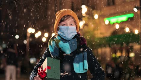 retrato de un niño caucásico feliz con bufanda sosteniendo un regalo y sonriendo a la cámara mientras nieva en la calle en navidad