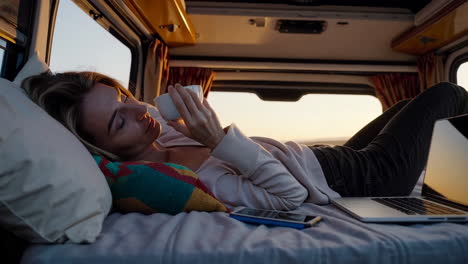 woman relaxing in a camper van with a cup and laptop at sunset or sunrise