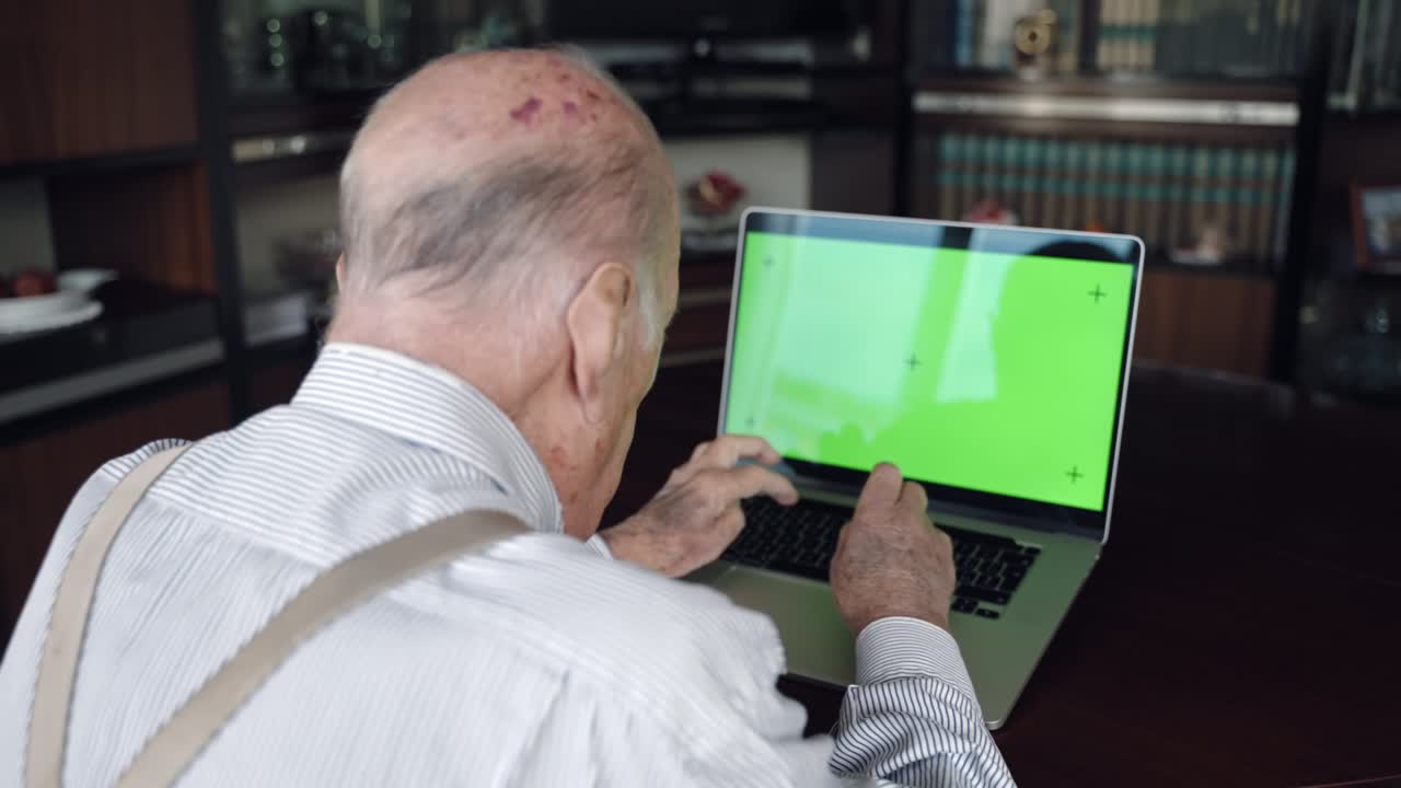 vista trasera de abuelo solitario sentado en la mesa de escritorio frente a la computadora de moda con pantalla verde croma maqueta de tecla, escribiendo en el teclado y haciendo llamada de video en línea con los miembros de la familia en