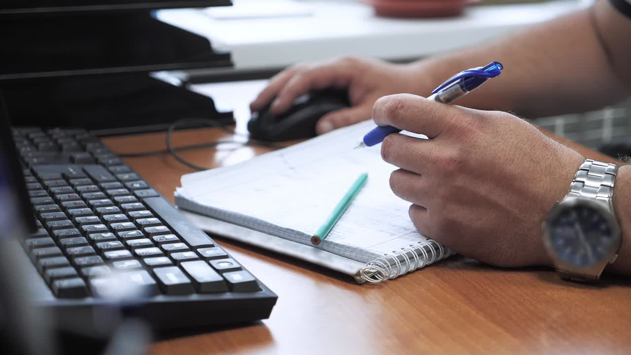 한 남자가 사무실에서 컴퓨터에서 일하고 노트북에 메모를 합니다.