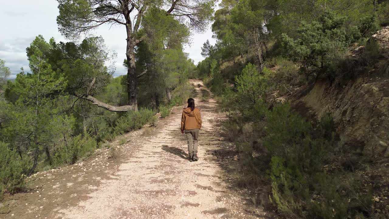 Brunette woman in a brown jacket and green pants walking on a leaf-covered forest trail, surrounded by pine trees and rosemary