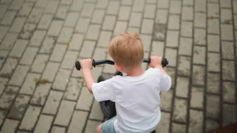 a child is seen from behind riding a small bicycle on an interlocked path, using his legs to propel the bike forward, the child is wearing a white t-shirt and shorts