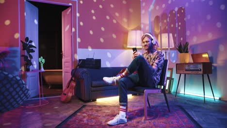 young stylish caucasian man with long hair in headphones and glasses sitting on a chair and using smartphone in a retro party at home